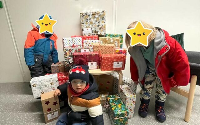 Kinder sitzen und stehen neben verpackten Weihnachtsgeschenken.